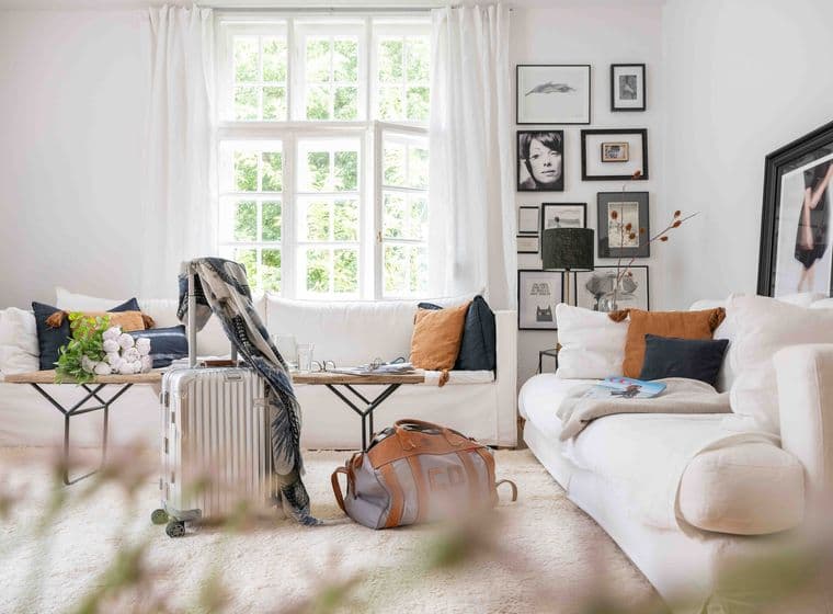 Cozy living room with white sofas, assorted pillows, a suitcase, and a travel bag. Large window and framed art on walls.