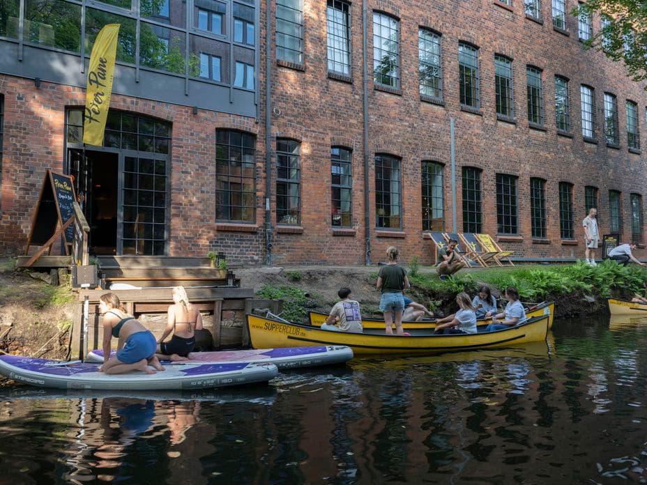 People relax on paddleboards and yellow boats by a brick building with large windows, next to a calm river.