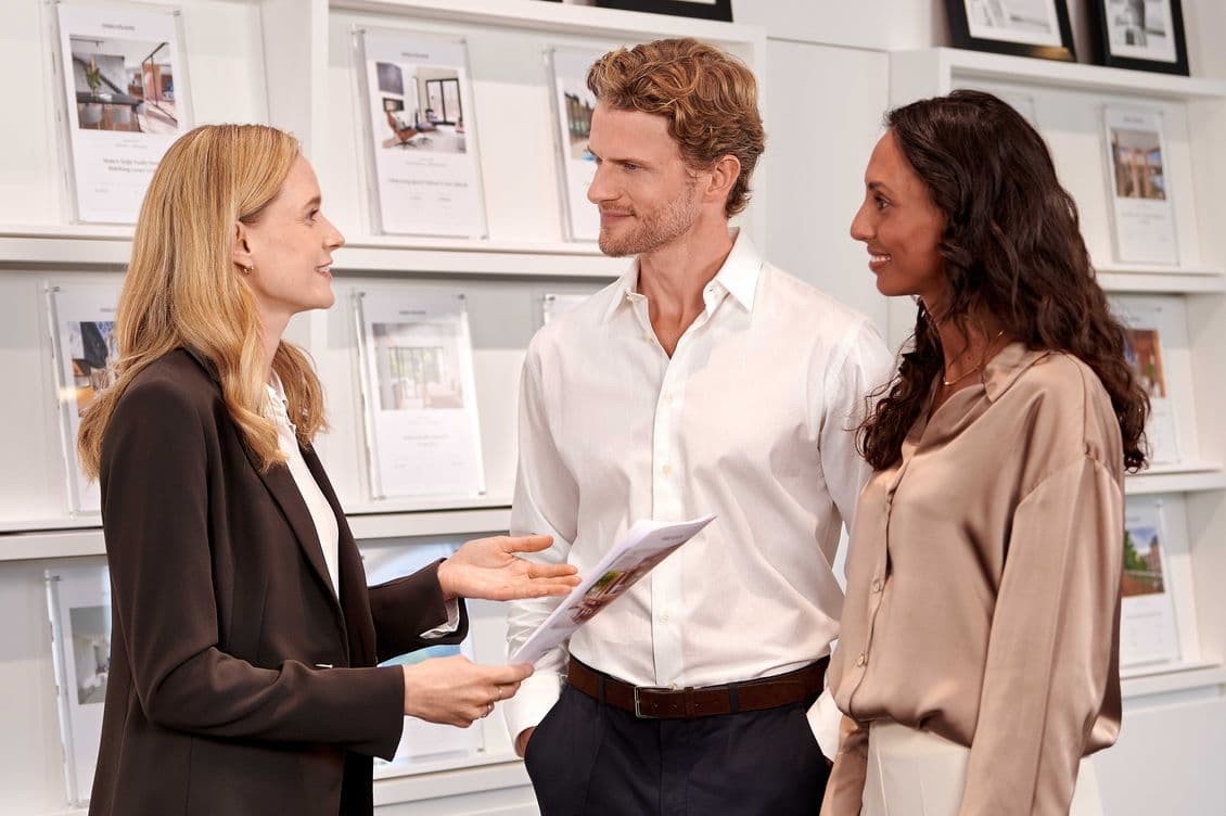 A real estate agent discusses property details with a smiling couple in an office, surrounded by framed pictures of homes on the wall.