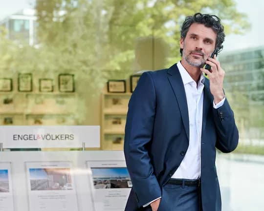 Man in a suit talking on a phone outside a real estate office with property listings visible through the window.