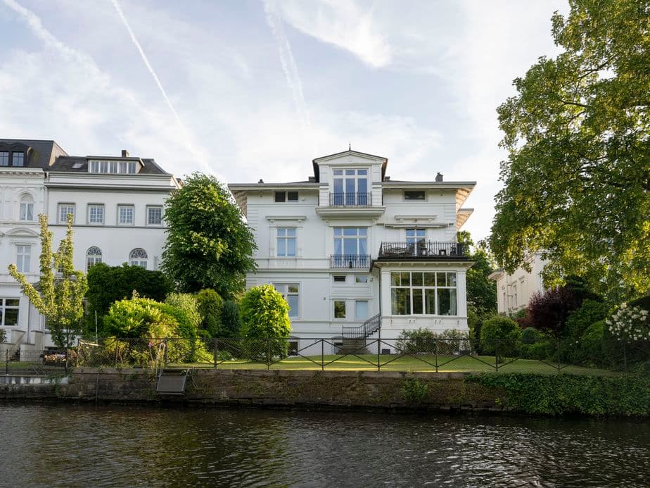 Elegant white house with large windows and a balcony, surrounded by lush greenery, situated on a quiet river under a partly cloudy sky.