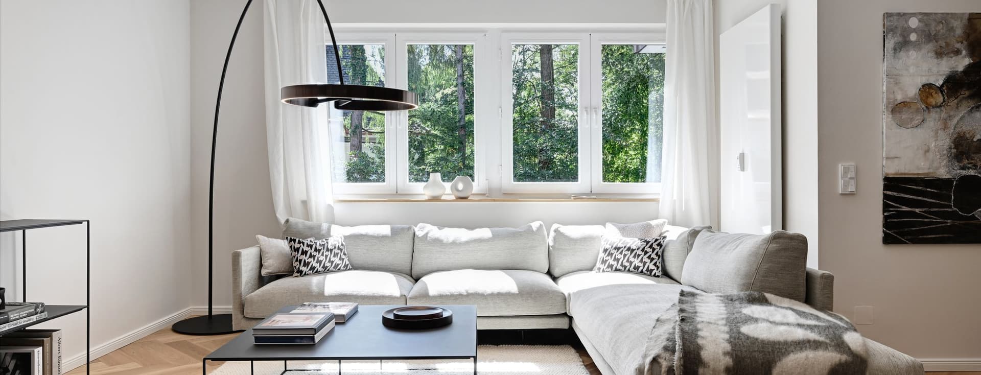 Modern living room with a large gray sectional sofa, black floor lamp, coffee table, and window with a view of greenery. Neutral decor and artwork.