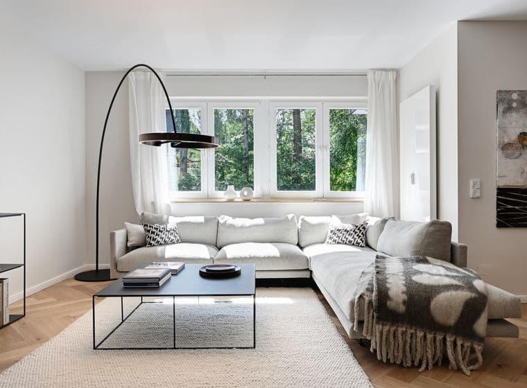 Modern living room with a large gray sectional sofa, black floor lamp, coffee table, and window with a view of greenery. Neutral decor and artwork.