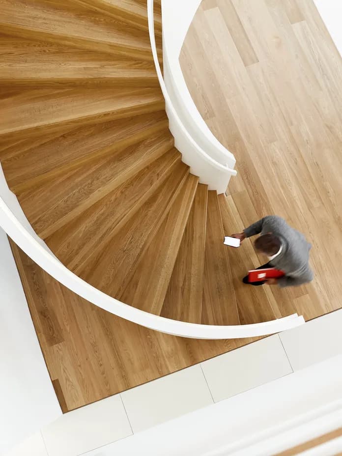 Bird's eye view of a modern spiral staircase with wooden treads and white curved balustrade, featuring a person walking up the stairs. The elegant design combines natural oak flooring with contemporary white architectural elements.