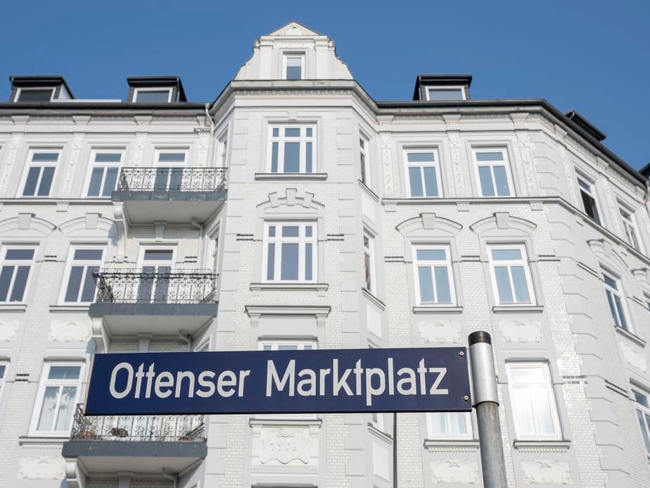 Street sign with the inscription ‘Ottensers Marktplatz’ in front of a white, multi-storey building with ornate architecture under a clear blue sky.