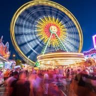 A vibrant fairground at night with a large, illuminated Ferris wheel in motion, surrounded by colorful stalls and a crowd of blurred people.