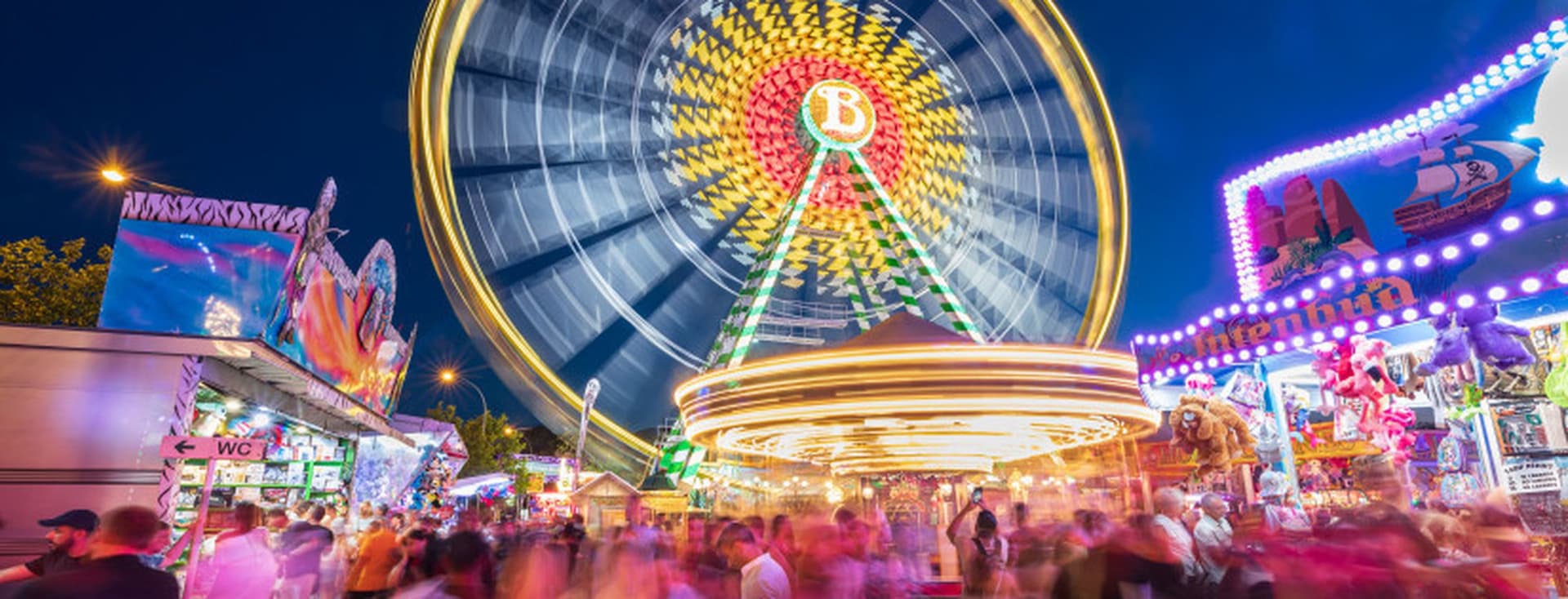 A vibrant fairground at night with a large, illuminated Ferris wheel in motion, surrounded by colorful stalls and a crowd of blurred people.