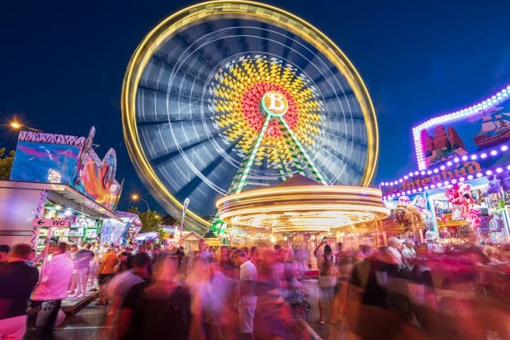 A vibrant fairground at night with a large, illuminated Ferris wheel in motion, surrounded by colorful stalls and a crowd of blurred people.