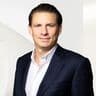 Sebastian König in einem dunklen Anzug und weißem Hemd vor einem hellen, abstrakten Hintergrund.