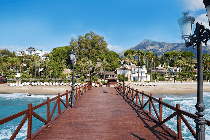 Bridge of the Marbella Club Hotel with lampposts on the sides, above the sea with waves, and in the background the Marbella Club Hotel