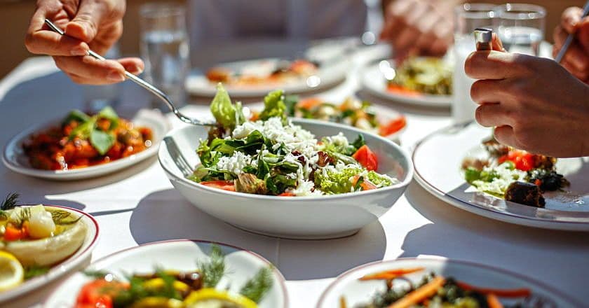 Se ven nueve platos de cocina mediterránea, ensaladas, y dos personas comiendo