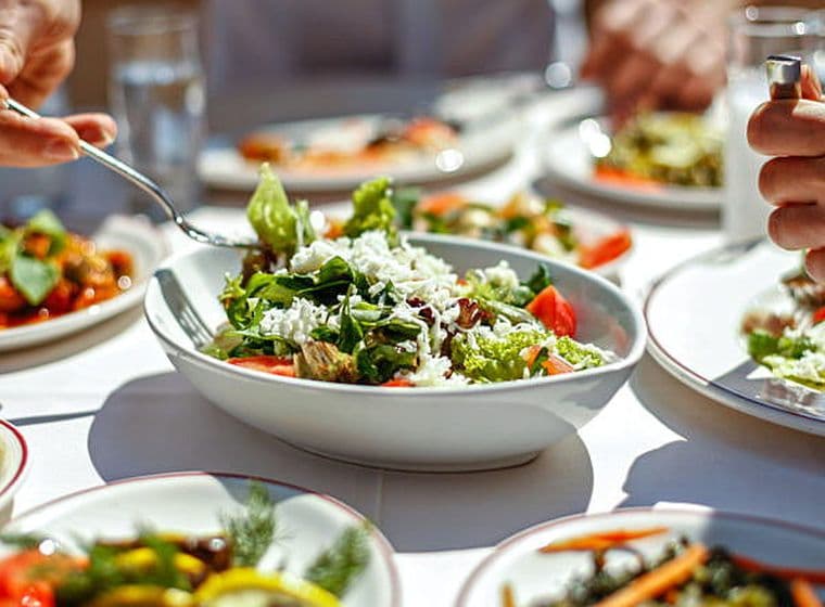 Es veuen nou plats de cuina mediterrània, amanides, i dues persones menjant.