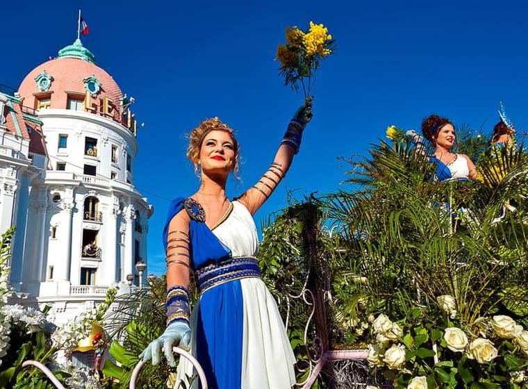 A woman in a blue and white dress holds yellow flowers at the Nice Flower Parade, with a building in the background.