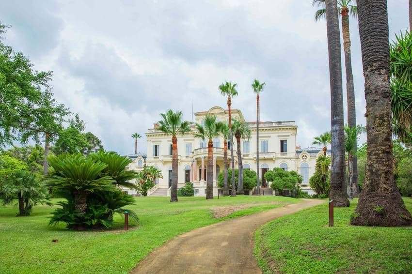 Élégante demeure couleur crème avec des palmiers et une pelouse verdoyante sous un ciel nuageux. Un chemin sinueux mène à la grande entrée.