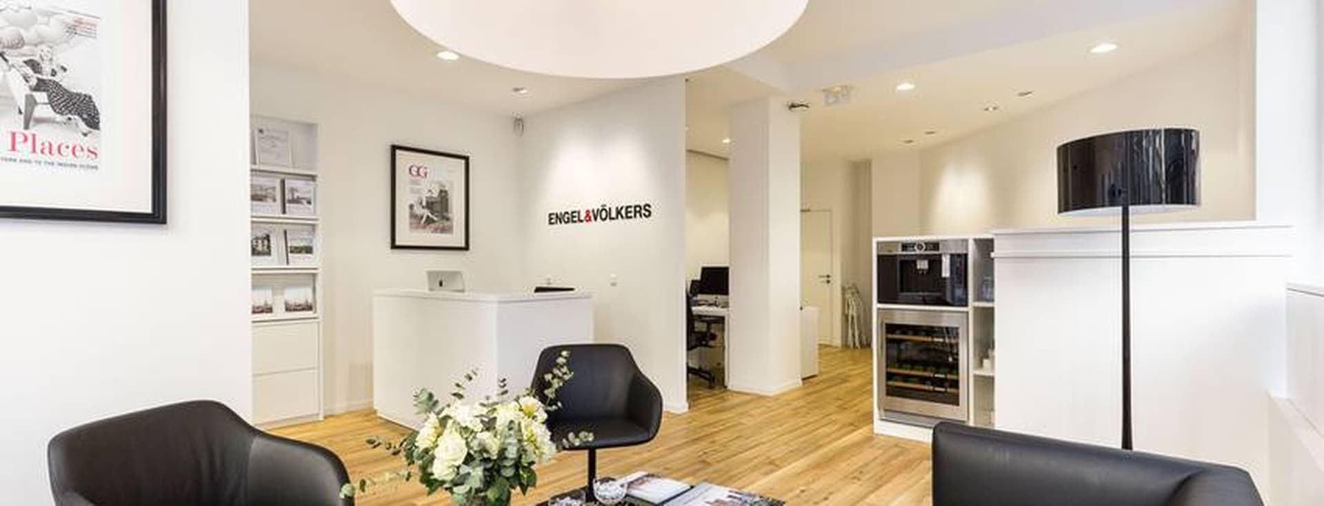 Hall d'entrée du bureau Engel & Völkers avec des sièges en cuir noir, une table basse noire avec des magazines et une réception blanche.