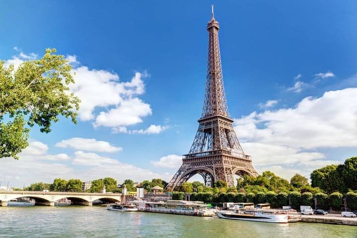 Eiffel Tower view from the Seine River, Paris. Boats docked along the riverbank, bridge in the background, blue sky with clouds.