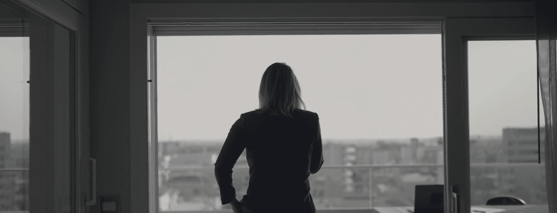 A woman in a blue suit looks out a window at a city skyline. A table and laptop are visible in the room.