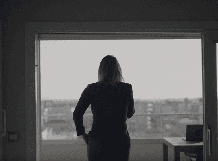 A woman in a blue suit looks out a window at a city skyline. A table and laptop are visible in the room.