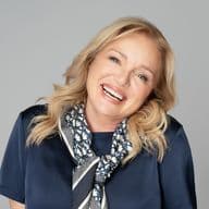 Smiling blonde woman in navy blouse and patterned scarf against a gray background.