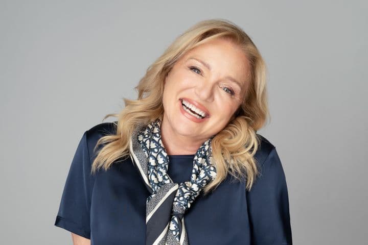 Lächelnde blonde Frau in marineblauer Bluse und gemustertem Schal vor einem grauen Hintergrund.