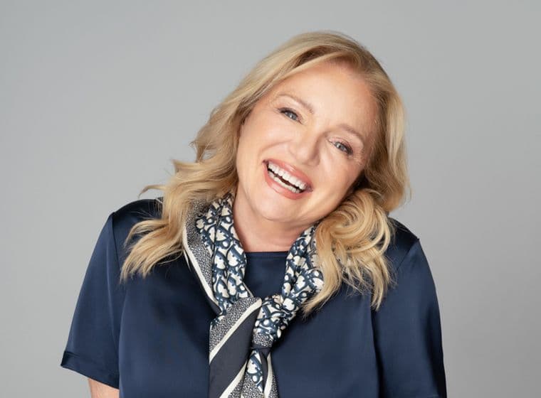 Lächelnde blonde Frau in marineblauer Bluse und gemustertem Schal vor einem grauen Hintergrund.