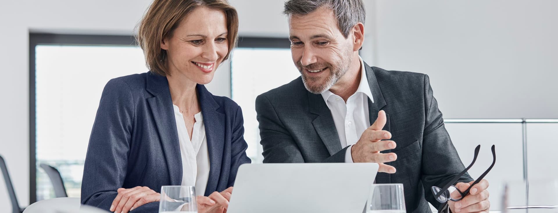 Zwei Fachleute in Geschäftskleidung diskutieren und lächeln, während sie in einer modernen Büroumgebung auf einen Laptop schauen.