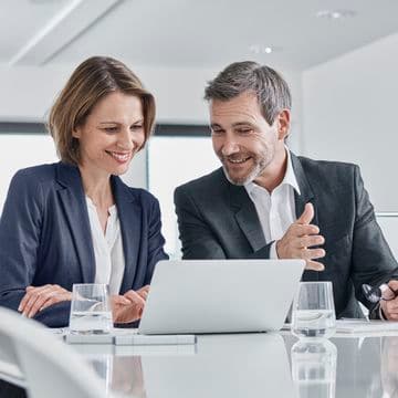Two professionals in business attire discussing and smiling while looking at a laptop in a modern office setting.
