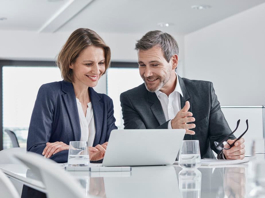 Two professionals in business attire discussing and smiling while looking at a laptop in a modern office setting.