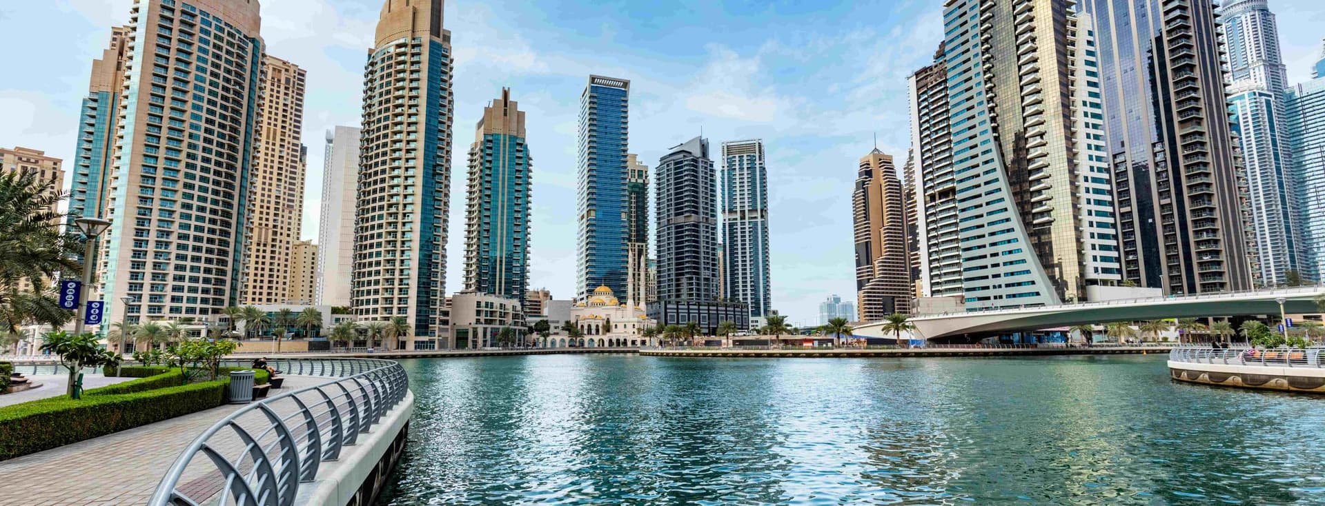 Modern skyscrapers line the waterfront of Dubai Marina, with a clear blue sky and calm water reflecting the buildings.