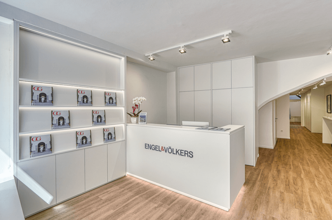 Modern real estate office with white walls, wooden flooring, a reception desk, and shelves displaying magazines. Bright and minimalist design.