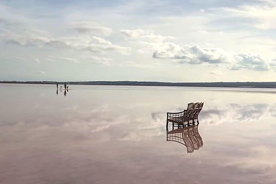 Agua rosada de la laguna con dos sillas puestas en el agua reflejando el cielo con nubes y personas andando en el horizonte