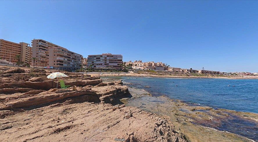 Vista de la Platja Gallina Torrevella amb les seves roques i edificis a l'esquerra.