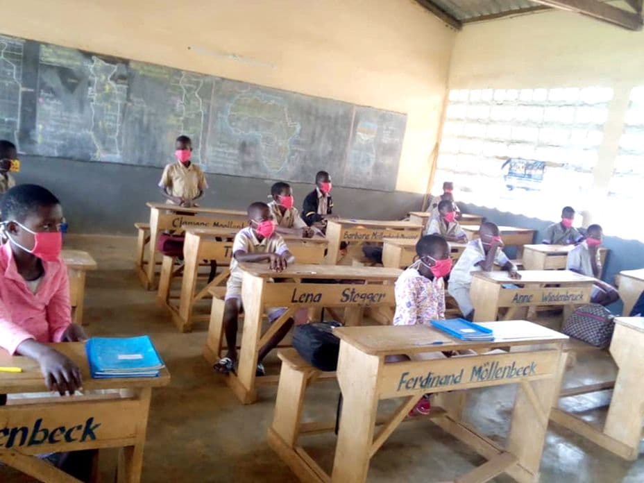 Students in a classroom, wearing red masks, seated at wooden desks with names. Chalkboards with writing are on the wall in the background.