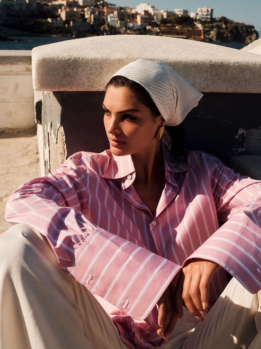 Μια γυναίκα με ροζ ριγέ πουκάμισο και λευκό παντελόνι κάθεται έξω με μια λευκή μαντίλα, κοιτάζοντας μακριά από την κάμερα. Μια παραθαλάσσια πόλη βρίσκεται στο βάθος.