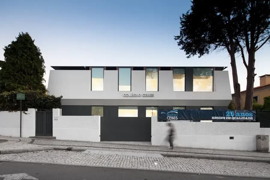 Ein modernes weißes Gebäude mit großen Fenstern, beschriftet mit "Colégio CEBES", hinter einer weißen Mauer und einem Tor. Eine Person geht auf dem gepflasterten Gehweg vorbei.