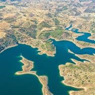 Luftaufnahme eines sich windenden blauen Stausees, umgeben von trockenem, hügeligem Gelände mit Flecken grüner Vegetation.