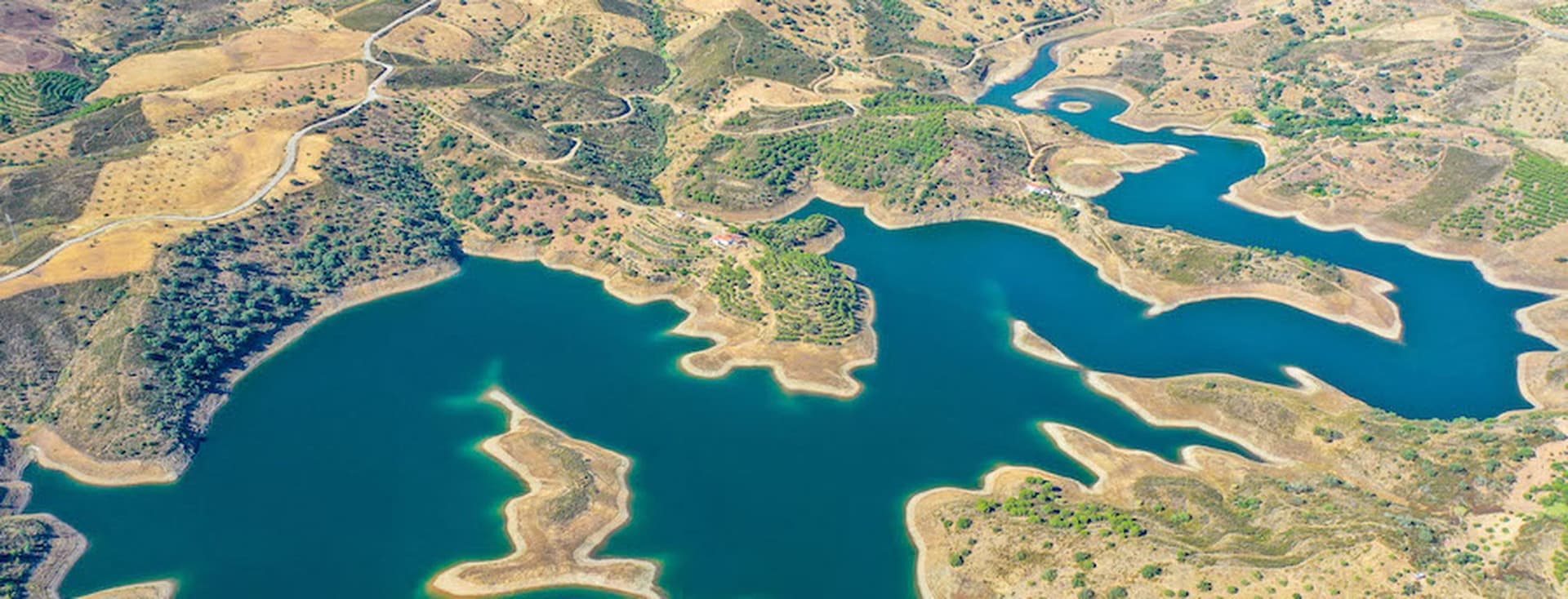 Aerial view of a winding blue reservoir surrounded by arid, hilly terrain with patches of green vegetation.