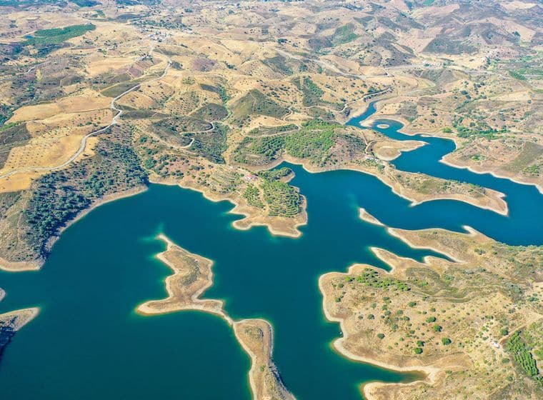 Luftaufnahme eines sich windenden blauen Stausees, umgeben von trockenem, hügeligem Gelände mit Flecken grüner Vegetation.