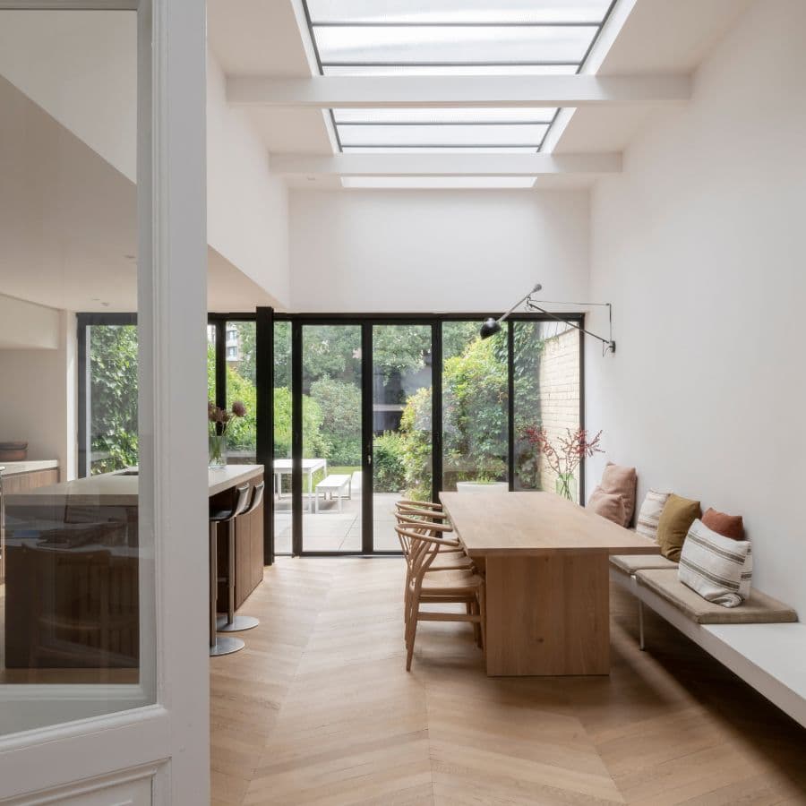Bright, modern dining area with wood table, chairs, and bench seating. Large glass doors open to a garden. Skylights illuminate the white walls and herringbone floor.