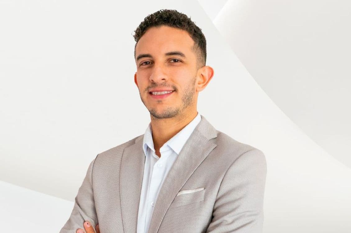 Un hombre sonriente con un traje gris está de pie con los brazos cruzados sobre un fondo blanco.