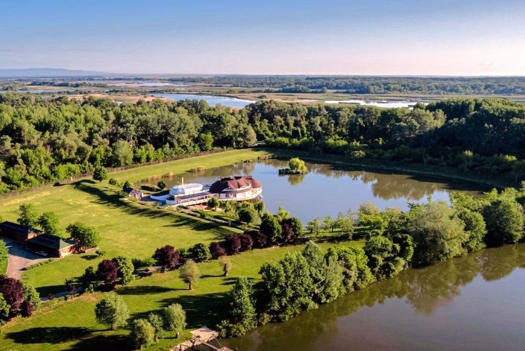 Aerial view of a lush green landscape with a large pond in Hungary, surrounding trees, and a circular building near the water's edge under a clear blue sky.