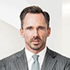 Benjamin Rogmans in a gray pinstripe suit with a white shirt and light gray tie, standing against a light background, looking directly at the camera.