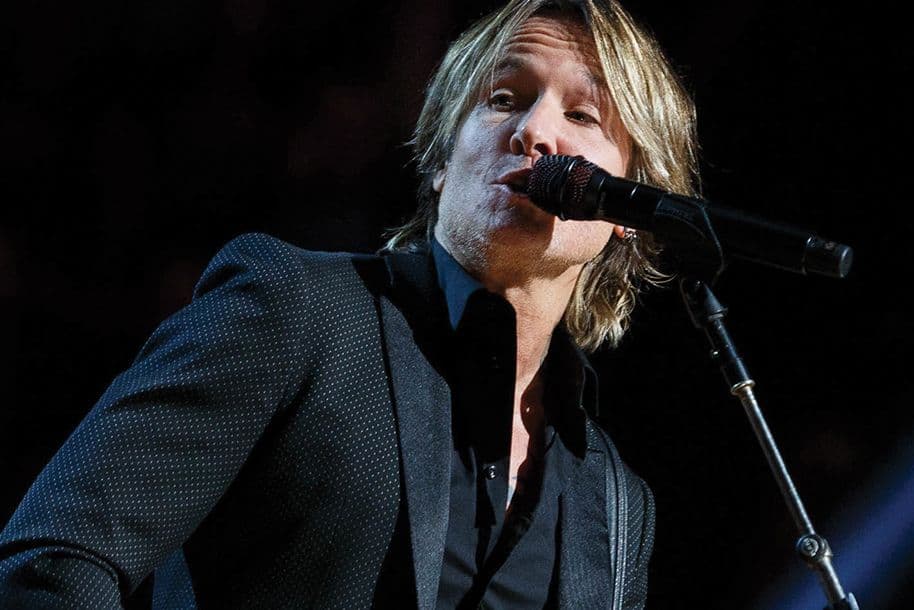 Keith Urban sings into a microphone. He wears a black suit and shirt. The background is dark.