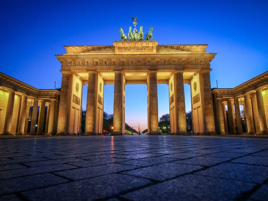 Brandenburg Gate at dusk, illuminated against a deep blue sky, with cobblestone pavement in the foreground.