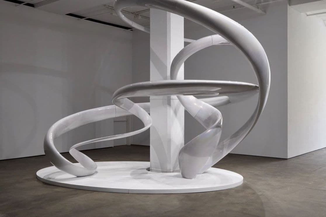 Weiße abstrakte Skulptur mit gewundenen spiralförmigen Bändern auf einem runden Sockel in einer modernen Galerie.