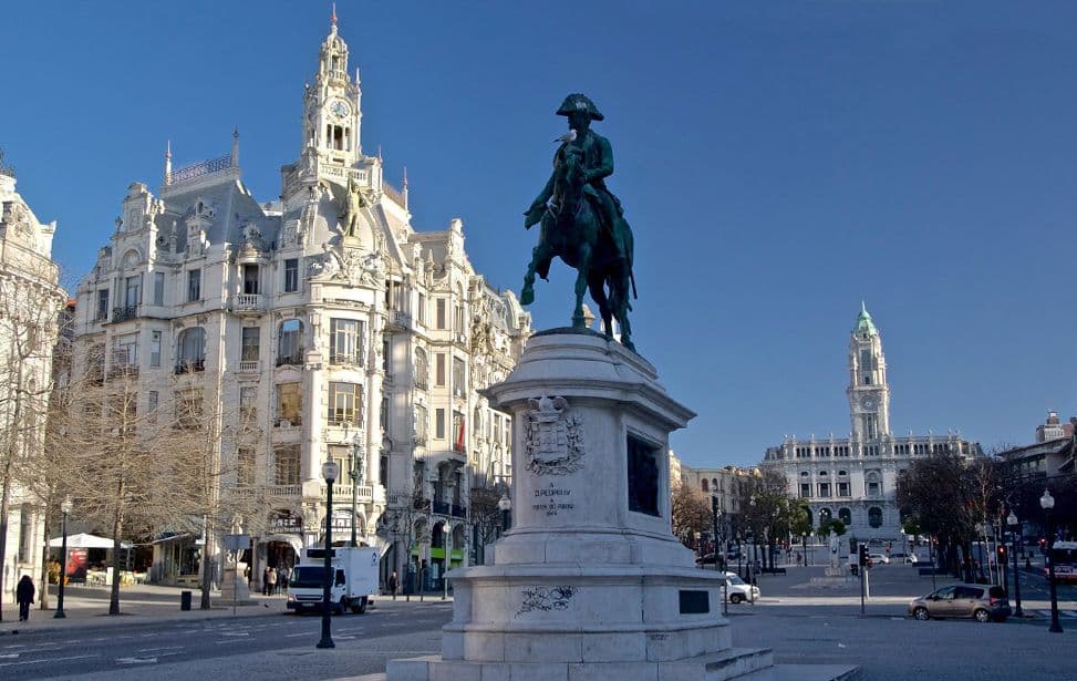 Statue of a rider on horseback in a plaza with ornate buildings and a clock tower under a clear blue sky.