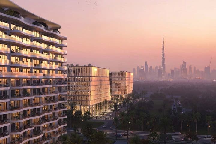 Sunset view of a curved glass-fronted residential tower with balconies, palm-lined streets, and a distant skyline topped by a tall spire.