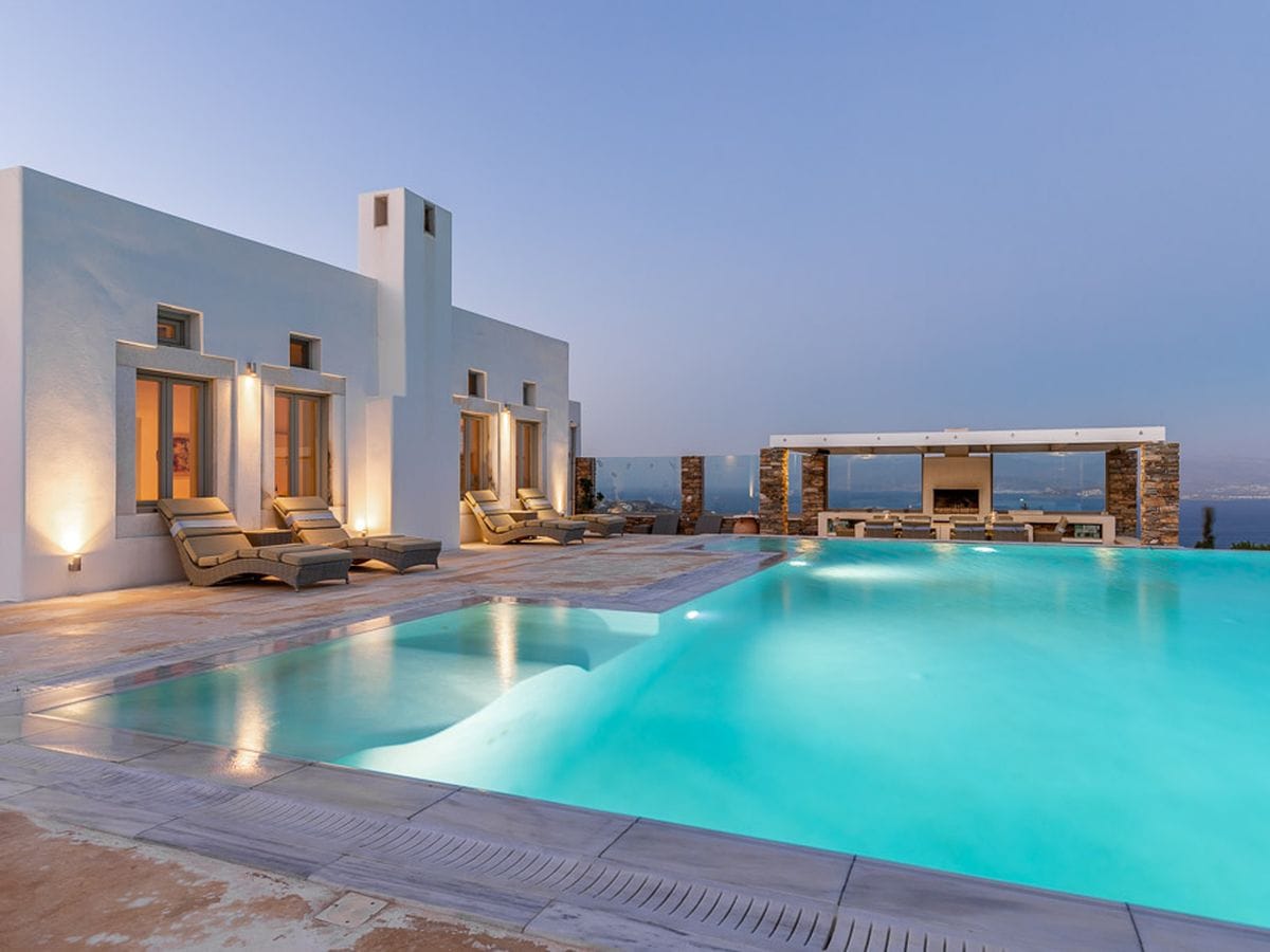 Luxury villa with a turquoise pool, lounge chairs, and an outdoor kitchen overlooking the ocean at dusk.