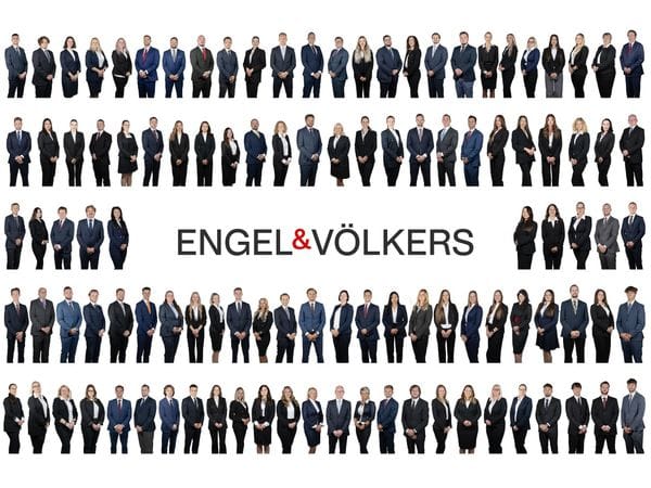 Group photo of the entire Engel & Völkers team. Dozens of men and women in business suits stand in several horizontal rows against a white background, with the Engel & Völkers logo prominently displayed in the center.