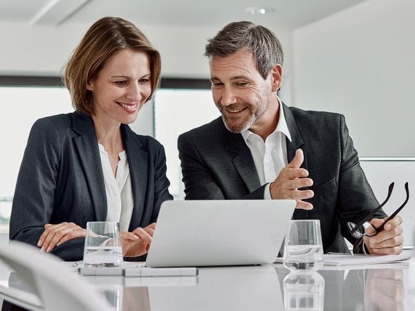 Two professionals in business attire, a woman and a man, sitting at a modern conference table reviewing content on a laptop. Both are smiling and engaged in discussion in a bright office setting with water glasses on the table.